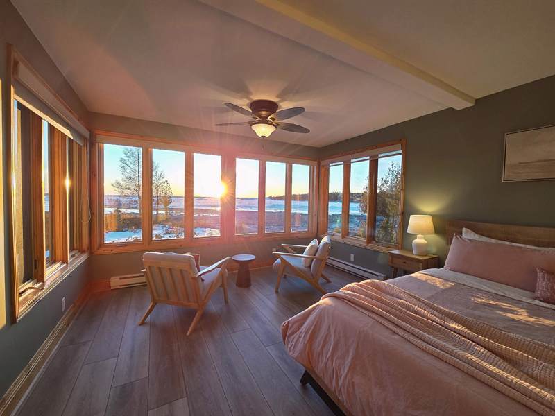 Bedroom at sunset with pink bedding, corner windows glowing over a snowy shoreline, two armchairs