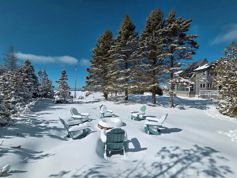 Teal Adirondack chairs and fire pit buried in deep snow, snowy spruces and the cottage at right