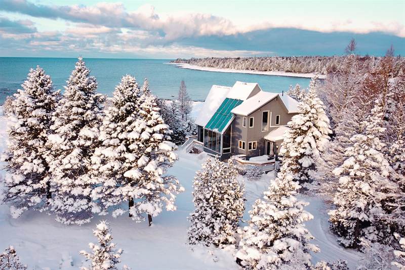 Aerial view of the cottage and snow-laden evergreens beside open water and a snowy curving shoreline