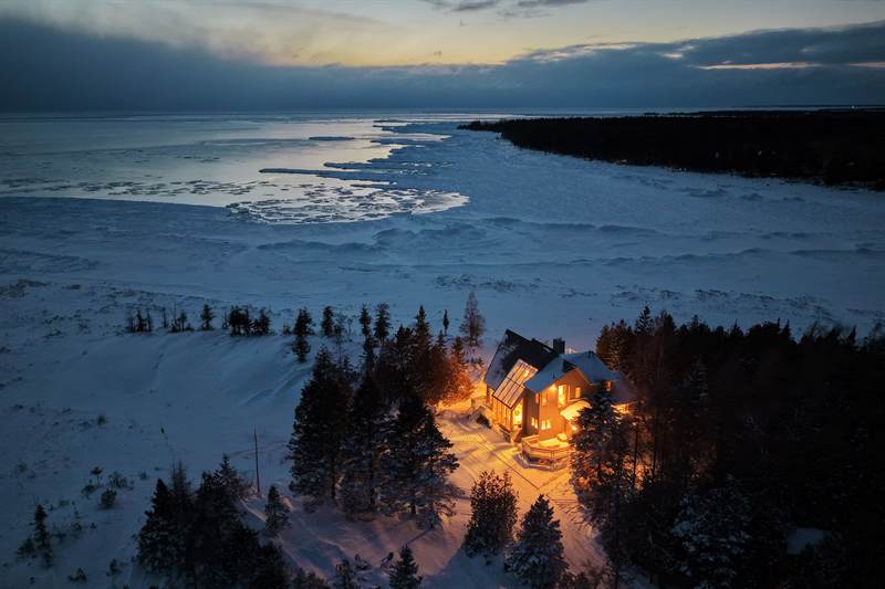 Winter dusk aerial of the glowing cottage by the frozen bay